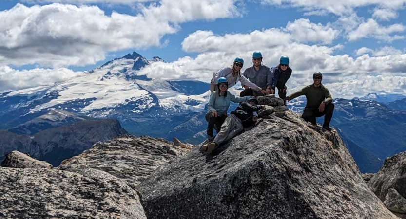 A group of people wearing helmets gather around a rocky summit. In the background there is a huge snow-covered mountain. 